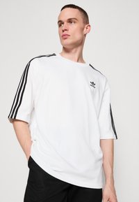 Balta trumpa rankovė t-tankas su trimis juodais dryžiais per pečius ir Adidas logotipu krūtinėje, pagamintas iš lygaus audinio.