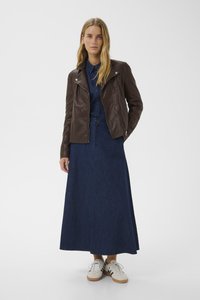 Giacca da biker in pelle marrone sopra un vestito di jeans blu con bottoni frontali, abbinato a sneakers bianche. Tessuto morbido, design aderente, lunghezza maxi.