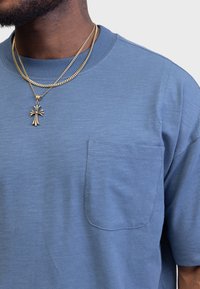 Blue short-sleeve T-shirt with a pocket on the left side, made from a soft, textured fabric. Accented with layered gold chain necklaces.