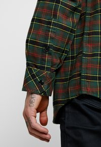 Hand med fingertatueringar under ärmslappen på en långärmad skjorta med grön, röd och gul rutigt mönster, bredvid svarta byxor.