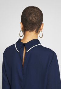 Granatowa bluzka z białym wykończeniem kołnierzyka, z wycięciem na plecach i gładką fakturą. Długie rękawy o luźnym kroju.