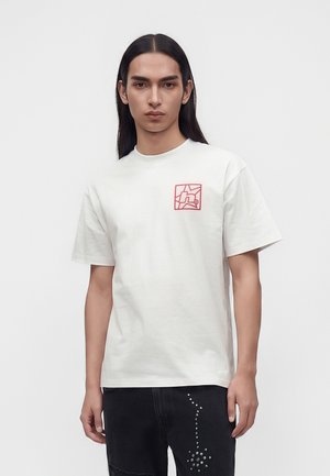 Ung person med langt mørkt hår bærer en hvid t-shirt med et lille rødt motiv på brystet og sorte bukser med nitte-mønster.