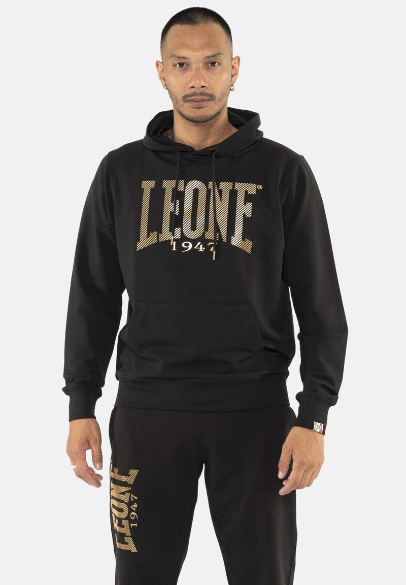 Homme portant un sweat à capuche noir et un pantalon avec le texte doré « LEONE 1947 », debout face à l’appareil devant un fond uni.