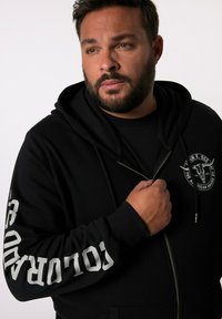 Sudadera con cremallera negra y un gran logo en el lado izquierdo del pecho, y texto en blanco en la manga derecha. Tela suave y capucha con cordones.