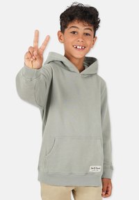 Ljusgrön hoodie i mjukt material, med en framficka, ribbade ärmslut och en etikett i nederkant. Ett barn gör tecknet för fred.
