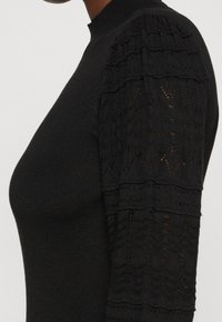 Sort trøje med en tætsiddende design, teksturerede strikkede ærmer med blonderlignende mønstre og høj lukning.