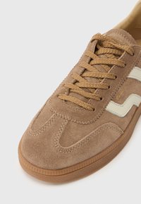 Ténis de camurça castanho claro com atacadores da mesma cor, logótipo lateral em creme e sola de borracha texturada sobre fundo branco.