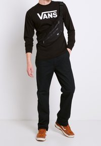 Mustapaitainen pitkähihainen paita, jossa on "VANS"-logo, musta olkalaukku, mustat housut ja ruskeat lenkkarit. Laukussa on vetoketjullisia taskuja.