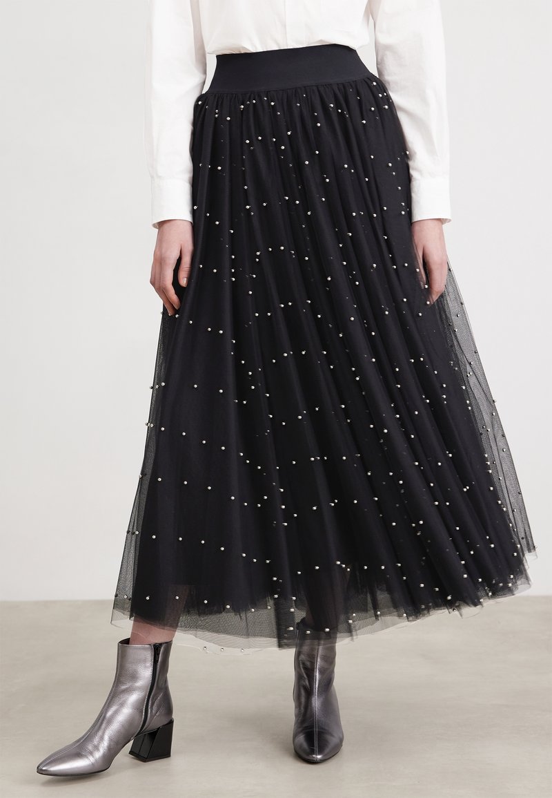 Femme portant une jupe midi en tulle noire ornée de petites perles blanches et des bottines métalliques argentées, associée à une chemise blanche à manches longues.