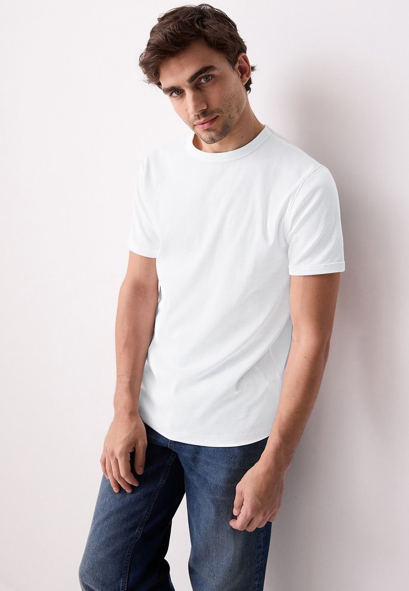 Jeune homme aux cheveux foncés portant un t-shirt blanc uni à manches courtes et un jean bleu, s'appuyant légèrement contre un mur blanc.