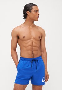 Shorts de bain bleus en tissu lisse avec une taille à cordon de serrage et un logo Nike blanc à l'avant. Conçus pour un ajustement ajusté.