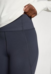 Hånd som justerer midjebåndet på høyhårede mørkegrå leggings med synlige sømmer, iført en hvit langermet topp.