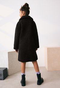 Manteau teddy noir en fausse fourrure avec col châle, coupe oversize, et manches longues, porté sur un pantalon longueur cheville et des bottes noires.