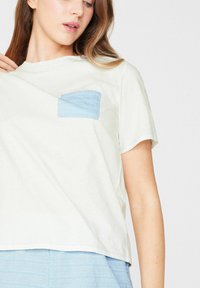 Joven mujer ajustando una camiseta blanca suelta con un bolsillo azul claro en el pecho, llevando pantalones azul claro a juego, sobre un fondo liso.