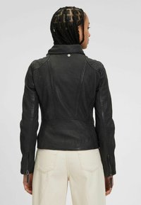 Giacca in pelle nera con finitura testurizzata, forma aderente e polsini con zip. Presenta un colletto a punta e dettagli sulle cuciture sulla parte posteriore.