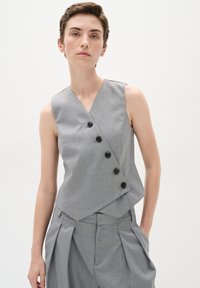 Grauste sleeveless Weste mit V-Ausschnitt, asymmetrischem Saum und fünf schwarzen Knöpfen. Hergestellt aus einem strukturierten Stoff, kombiniert mit passenden Hosen.