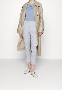 Beige läder trenchcoat över en ljusblå skjorta, matchat med randiga cropped byxor och vita loafers med guldaccent.