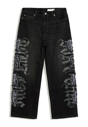 Jean en denim noir à jambes larges, avec des patchs de lettres de style gothique blanc et gris sur les deux jambes du pantalon.