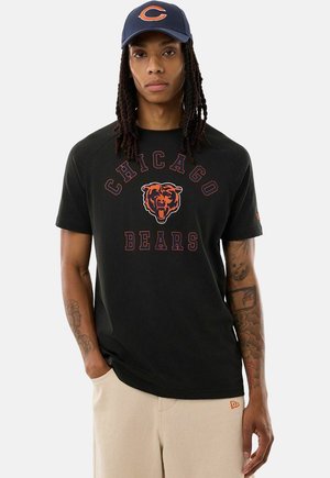 Persoon met lange dreadlocks, die een marineblauwe pet en een zwart Chicago Bears T-shirt draagt, staand met één hand in de zak van beige broek.