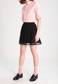 Kvinna iklädd en rosa skjorta med knappar, svart knälång kjol, rosa strumpor med ränder och svarta loafers, som står mot en enfärgad bakgrund.