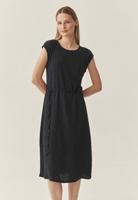 Crna haljina do koljena napravljena od teksturiranog materijala, s kratkim rukavima i pojasom s vezicom. Jednostavan, ravni kroj s suptilnim detaljima.