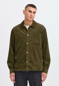 Chemise en velours côtelé vert avec col, boutons sur le devant et une poche poitrine unique. Manches longues avec poignets boutonés, texture côtelée discrète.
