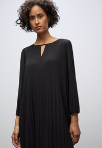 Schwarzes Kleid mit vertikalen Falten, runder Ausschnitt mit einem Schlüssellochausschnitt und drei Viertel-Ärmel mit engen Manschetten. Glatte Textur.