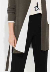 Cardigan vert olive avec doublure intérieure blanche, manches longues, devant ouvert, poches latérales et ceinture en tissu. Associé à un pantalon noir.