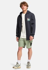 Sudadera con cremallera en color marino, con cordones y un gráfico en el pecho, combinada con pantalones cortos verde claro que cuentan con un bolsillo tipo cargo y detalles de malla.