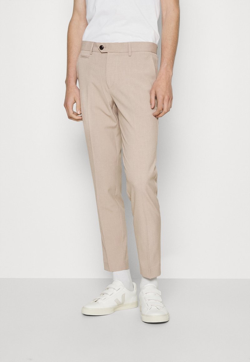 Homme portant un pantalon beige slim, des chaussettes blanches, des baskets blanches et un t-shirt blanc, debout devant un fond uni.