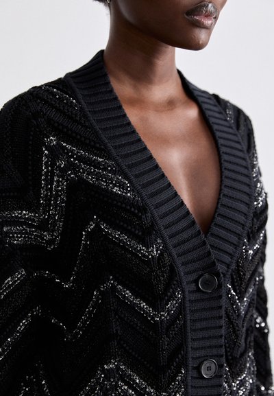 Μαύρο κασκόλ με υφή ζιγκ-ζαγκ και πούλιες. Ριπ επενδύσεις και δύο μαύρα κουμπιά μπροστά.