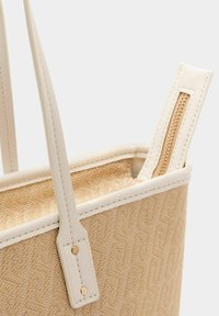 Beige tote bag met een textuur van geweven oppervlak en witte leren accenten, voorzien van een ritssluiting en stevige schouderbanden.