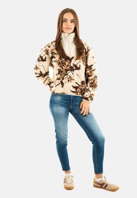 Giacca in pile a fiori crema con colletto alto, abbinata a jeans blu aderenti e sneakers beige con strisce bordeaux.