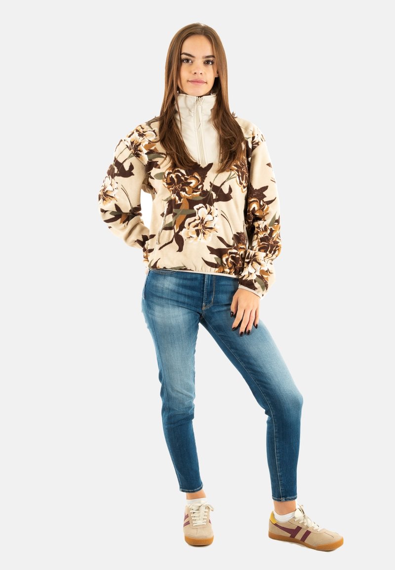 Giacca in pile a fiori crema con colletto alto, abbinata a jeans blu aderenti e sneakers beige con strisce bordeaux.