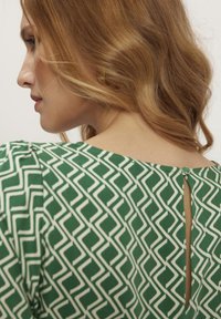 Zielona sukienka w geometricalne wzory z białymi detalami, z rozcięciem w kształcie klucza na plecach i małym guzikiem, wykonana z gładkiego materiału o miękkim układzie.