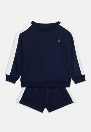 Survêtement pour enfants bleu marine avec haut à manches longues et short assorti, tous deux ornés de bandes latérales blanches et d’un petit écusson doré en forme d’ours sur la poitrine.