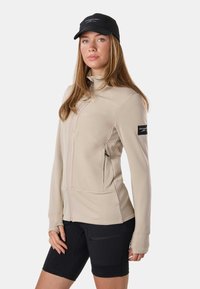 Femme aux longs cheveux portant une veste beige à fermeture éclair, un short noir avec une poche zippée, et une casquette noire avec un logo blanc.