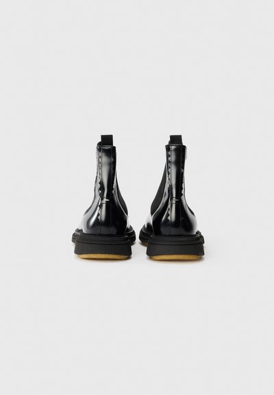 Botines de charol negro con tacón alto, que presentan paneles laterales elásticos y una suela de goma texturizada. Diseño minimalista y líneas limpias.
