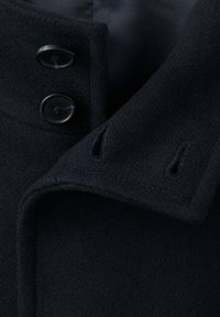 Granatowy kołnierz wełnianego płaszcza z dwoma czarnymi guzikami, wykonany z teksturowanej tkaniny z dziurkami na guziki, z widoczną miękką podszywką wewnętrzną.