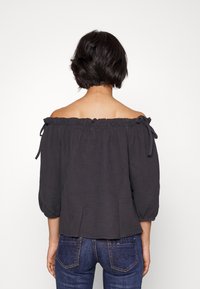 Off-shoulder blus i mörkgrått texturerat tyg, med volangkrage och knytband, i kombination med tajta blå jeans.