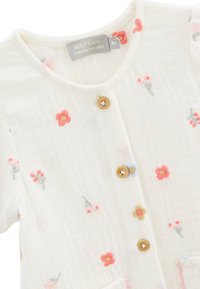 Cardigan en coton blanc avec broderies florales en rose, rouge et bleu. Doté de boutons en bois et de poches, il présente une texture douce et un design subtil.