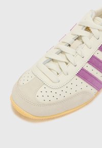 Vit sneaker med perforerat läder, lila mockastripes och sula av gummi. Snörningen är ljusgrå med sömdekoration.