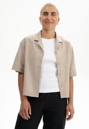 Person mit kurzen blonden Haaren trägt beige kurzärmliges Hemd über weißem Oberteil und schwarze Hose, steht vor weißem Hintergrund.