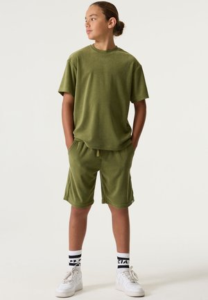 Giovane con i capelli raccolti in uno chignon, indossa una maglietta a maniche corte verde oliva e pantaloncini abbinati, scarpe da ginnastica bianche, in piedi con le mani nelle tasche.