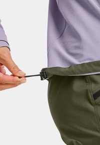 Elastic waist adjustment on olive green pants, with a hand pulling a black toggle. Light purple top partially visible above the waistband.