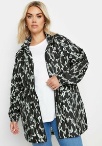 Svart och vit mönstrad lättviktsjacka med huva, dragkedja framtill och elastiska ärmmuddar, bärs över en vit t-shirt och blå jeans.