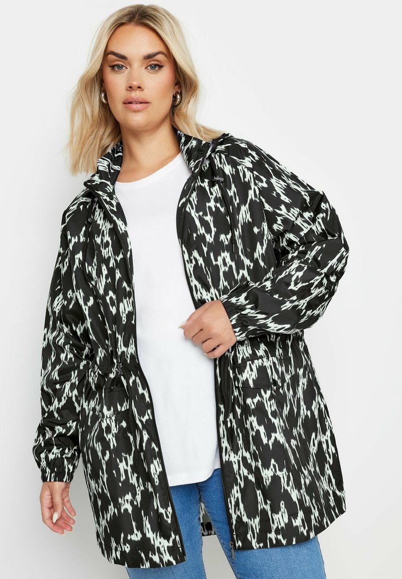 Svart och vit mönstrad lättviktsjacka med huva, dragkedja framtill och elastiska ärmmuddar, bärs över en vit t-shirt och blå jeans.