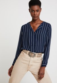 Marineblaues Langarmshirt mit weißen vertikalen Streifen, V-Ausschnitt, kombiniert mit hellbeigen Jeans und einem hellen Gürtel mit quadratischer Schnalle.
