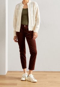 Osoba oblečená v béžovém cardiganovém svetru s otevřeným pletením, olivově zeleném tričku, vínových kalhotách a bílých teniskách stojící na dřevěné podlaze proti bílé zdi.