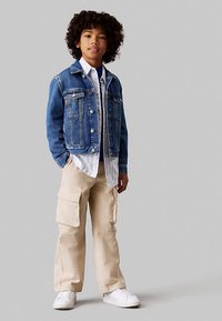 Jeans jakna svetlo modre prane barve, bela majica z dolgimi rokavi, bež cargo hlače z bočnimi žepi in beli supergi. Urbano priložnostno oblačilo.